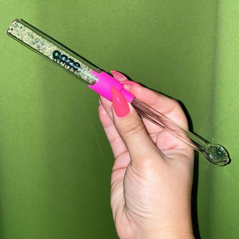 A white girl's hand with bright pink nails is holding a fully packed Ooze Glass Blunt Slider in front of a light green wall.