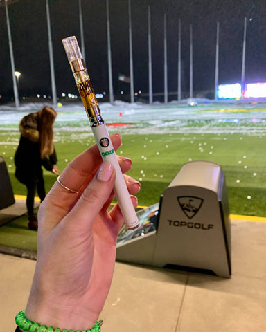 A white woman's hand is holding up a white Ooze Slim Twist Vape Pen with an oil cartridge attached while watching a friend take a swing at Top Golf.