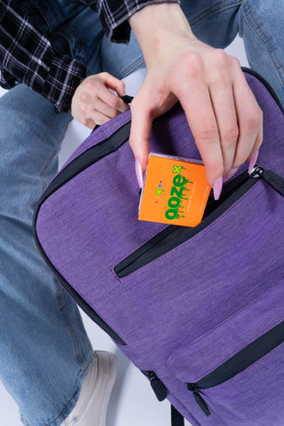 A girl is putting an orange OozeX box of concentrates into the front top pocket of a purple Ooze smell proof backpack