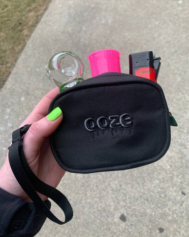 A smell proof black Ooze Wristlet is being carried outside, and is unzipped to show that it contains a hand pipe, pop top vial, and a lighter.