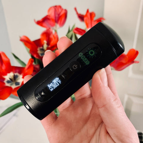 A black Ooze Drought dry herb vaporizer is turned on and held up in front of a white table with a vase full of red flowers.