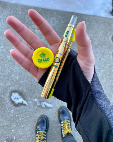 A white girl is standing outside, wearing Dr. Martens boots with yellow laces and holding the gold Ooze Fusion wax vaporizer, dab tool, and yellow stash jar.