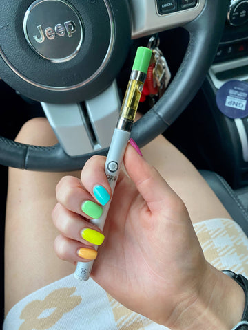 A white girl's hand with bright nails is holding the Ooze Polar Pearl Twist Slim Pen 2.0 with an OozeX cartridge attached. She's in her car.