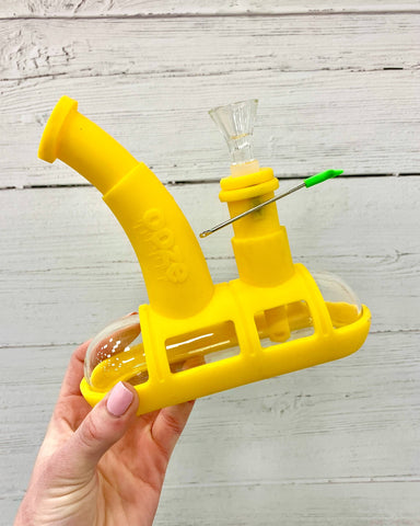 The yellow Ooze Steamboat is being held up in front of a white wood background. There is a small dab tool attached to the downstem by a magnet.