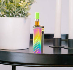 A rainbow Ooze Novex 2 vape has a cartridge inserted, and is sitting on a black coffee table next to a plant