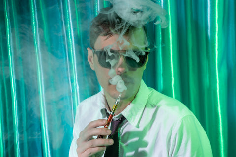 A white guy with glasses is exhaling vapor from a yellow Ooze Twist Slim Pen in front of a shiny teal curtain.