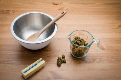 Preparing to make cannabutter. A silver mixing bowl is shown with a wooden spoon resting inside, there is a glass measuring cup filled with cannabis nugs, a few spare nugs on the wooden table, and a full stick of butter in the wrapper.