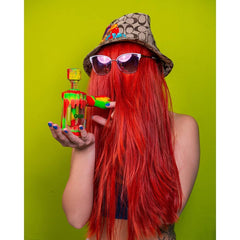 A girl with a long red wig covering her entire face is wearing a Coach bucket hat and sunglasses. She is holding a Rasta Ooze Clobb bong up in front of her face.