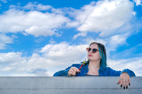 A young white woman with blue hair is looking over a gray wall with a blue sky and clouds in the background. She is holding a new Ooze Twist Slim Pen 2.0 up to her mouth.