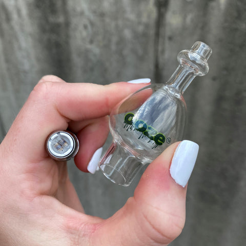 A girl is holding a vape battery with a coil attached and pinching a cloud glass globe with that same pointer and thumb.