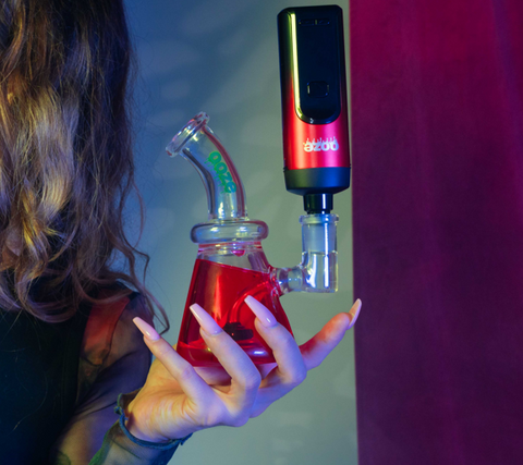 A girl with long pink acrylic nails is holding a red Ooze Glyco freeze bong that has a red and black Ooze Verge dry herb vape turned upside down and inserted into the bong