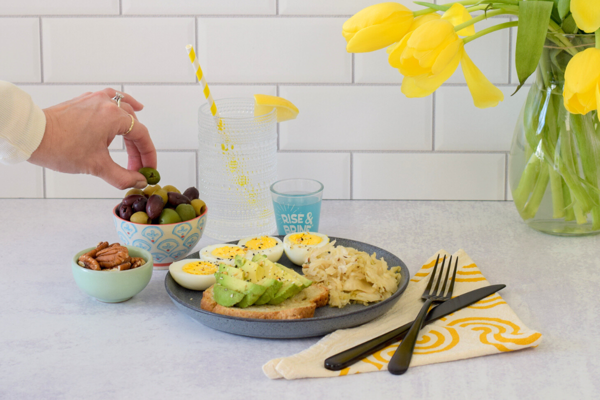A plate of avocado toast, boiled eggs, sauerkraut and a hand picking up an olive from a bowl.