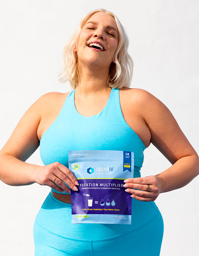 Woman in blue sports bra and leggings holding a 14ct pouch of Hydration Multiplier+ Probiotic Kombucha