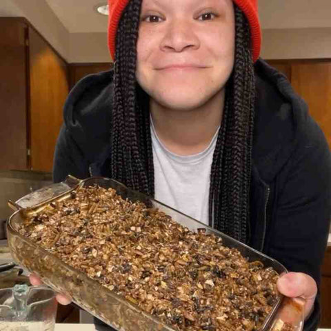 A glass pan of baked treats for chickens being held by a woman with long black braids and red beanie.