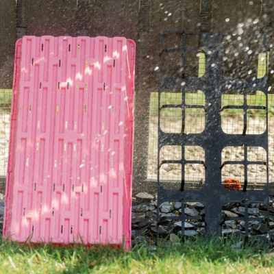 A Pink 1020 Tray with Holes Restings Against a Fence next to a 5" Pot Insert Holder being sprayed by a hose in a backyard.