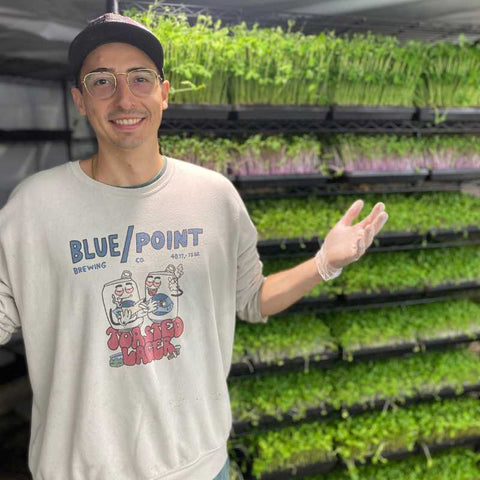 Donny Greens Standing near a tall rack of grown microgreens in 1020 trays