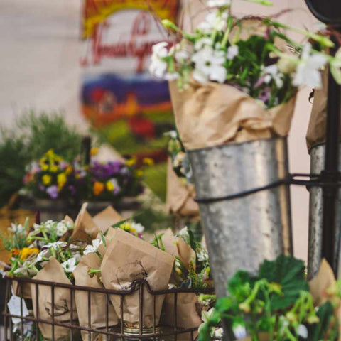 Bouquets of flowers wrapped in brown paper and placed in mason jars. The jarred bouquets are inside of a wire basket sitting on a curb.
