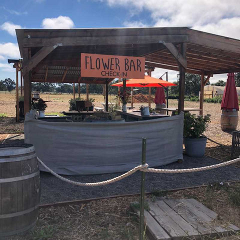 Wood and metal shade structure with an orange sign that reads "Flower Bar Check-in."