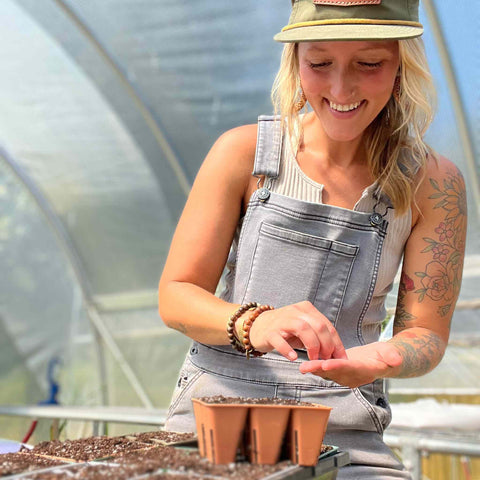 Blonde woman with hat starting seeds using a brown 6 cell plug tray in a greenhouse.