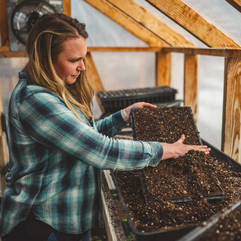 Female farmer in a flannel shirt filling a cell tray with seed starting mix inside a greenhouse.