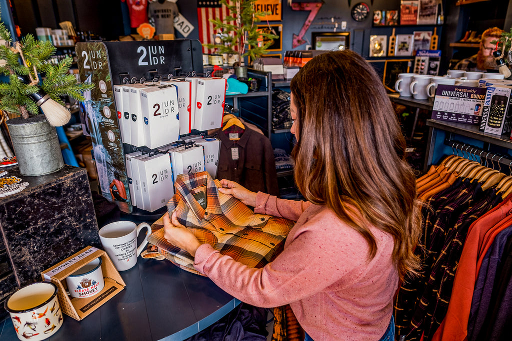 A mid-forties woman wearing a pink long-sleeved top is shopping at a men's store in Omaha. She is holding a flannel shirt from Howler Brothers Clothing.