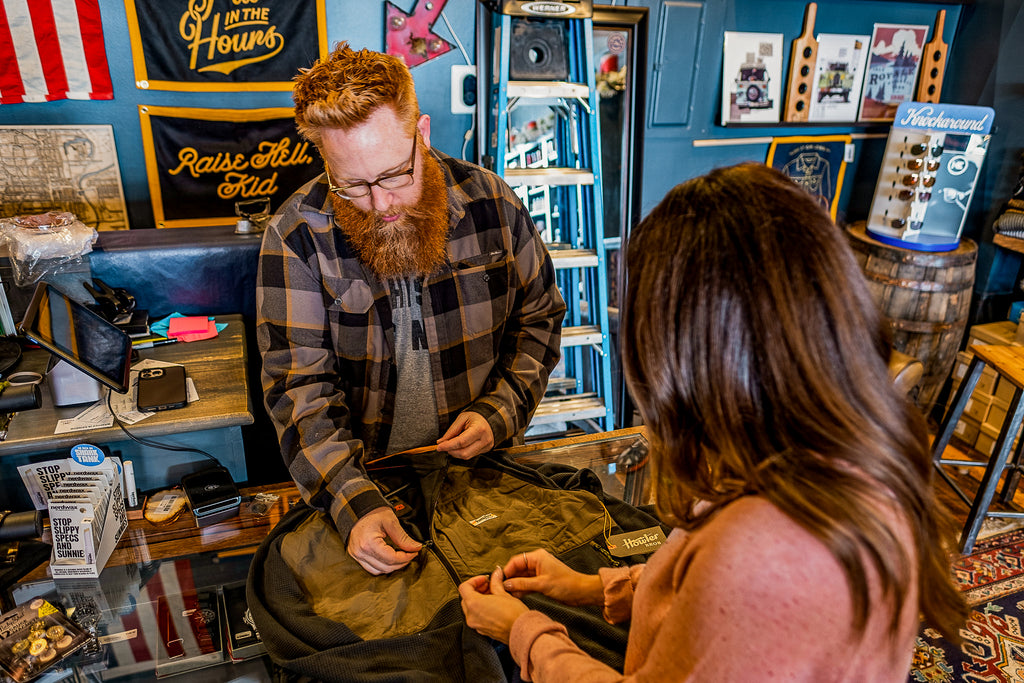 a man with a red beard and glasses is wearing a flannel shirt from Howler Bros. He is helping a young women pick out a long-sleeved jacket from Howler Brothers. 