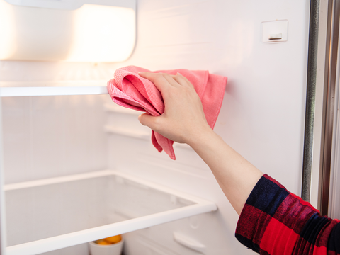 Inside of refrigerator featuring white interior and glass shelves. A hand holds a pink rag, cleaning the inside of the fridge.