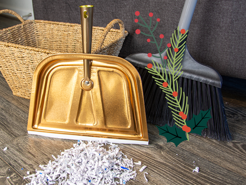 A copper motif dust pan sits behind a pile of shreded paper on a wooden floor. In the background there are holiday accents featuring holly berries and pine.