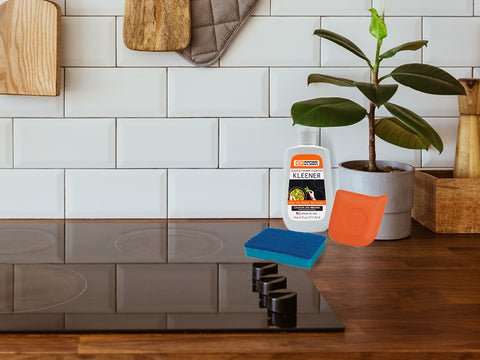 Image of kitchen glass cooktop with wooden countertops and white subway tile backsplash. Small bottle of cleaner, sponge and orange plastic scraper sit on top of the glass cooktop. Small plant in background.