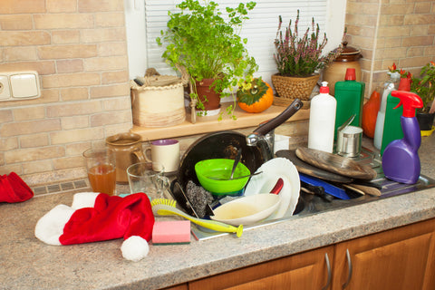 Beige and cream colored backsplash and countertop surround a kitchen sink overflowing with dirty dishes. Bottles of cleaner and plants are off to the side and back of photo. 