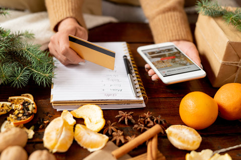 Person holds smart phone in one hand and credit card in another hand, a journal sits on a counter below featuring a list of indescript written list