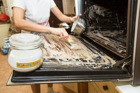 Image of a person cleaning a messy oven, brushing a baking soda paste onto the oven door while holding a stainless steel bowl. The oven has been brushed with paste. 