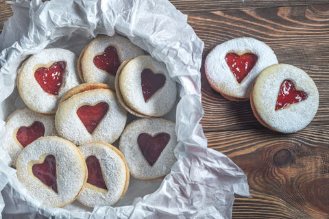 Round cookies dusted with confectioners sugar feature a heart in the center of each cookie filled with red jam.