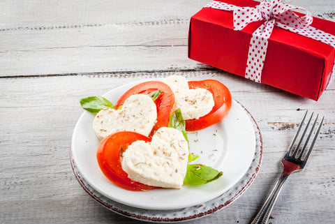 A caprese salad with heart shaped cheese, tomatoes and basil sit on a white plate.
