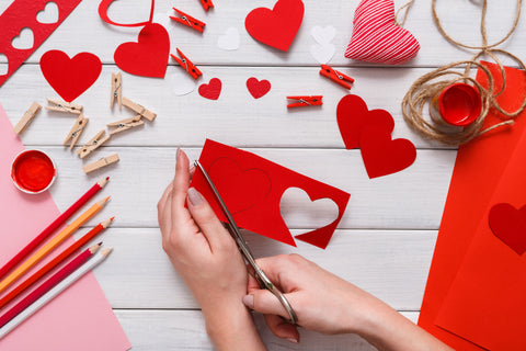White table background featuring hands cutting a heart out of red paper. Red paper, pencils and other art supplies sit on the table.