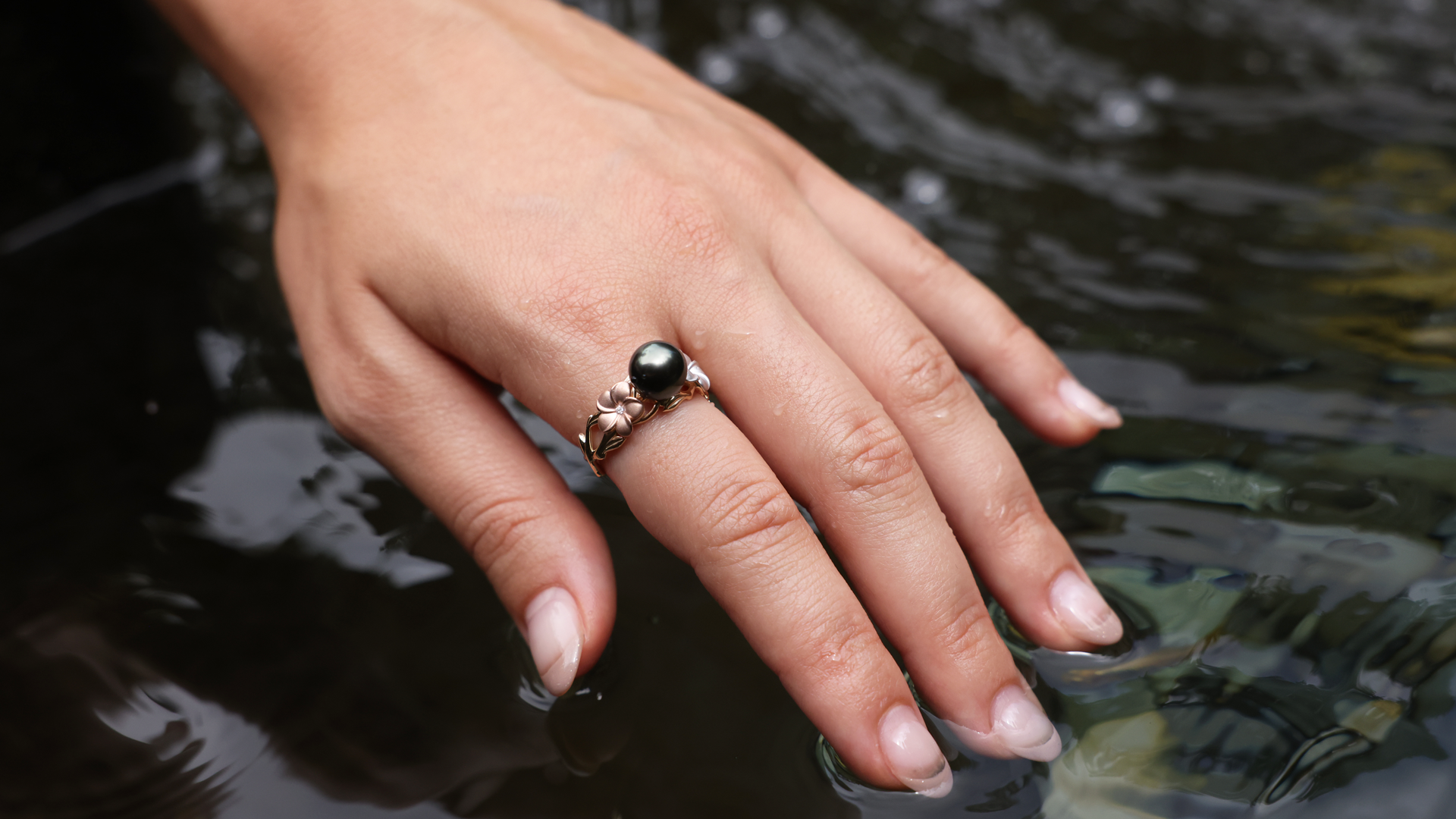 Main d'une femme avec une perle noire de Tahiti avec un anneau de Plumeria touchant l'eau