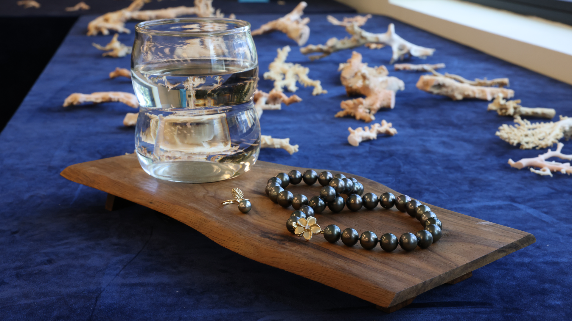 Ein Tahiti-Perlen- und Monstera-Ring, ein Tahiti-Schwarzperlenstrang mit Plumeria und ein Glas Wasser auf einem Tisch mit rosa Korallenstücken im Hintergrund