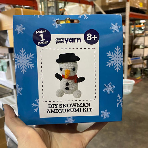 A hand is holding a blue snowman amigurumi kit in a warehouse