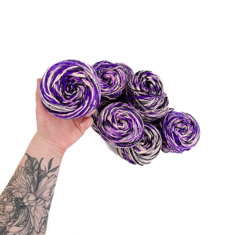 A tattooed hand is holding onto a skein of purple and white frostbite yarn, behind them a pile of frostbite worsted weight silk yarn