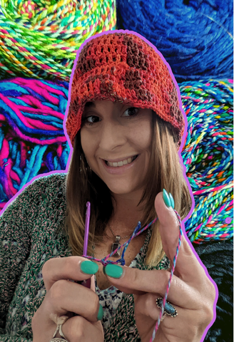 Jordan, a DGY worker with long brown hair and wearing a red hat is crocheting. Behind her are a stack of up close cakes of yarn.