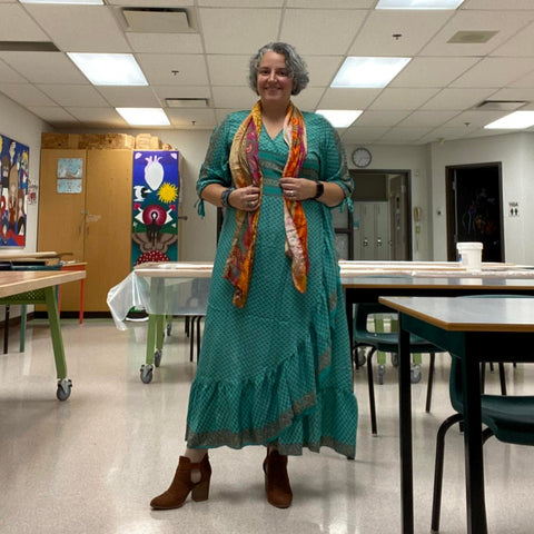 A woman with short grey hair is posing in a classroom, wearing a teal zaria dress, an orange scarf, and brown boots