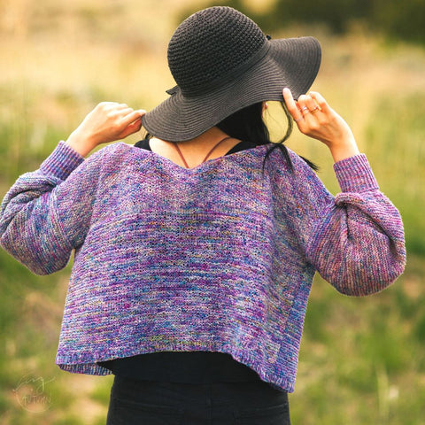 A person with a large black hat is facing away from the camera, wearing a purple and blue lace weight cardigan.