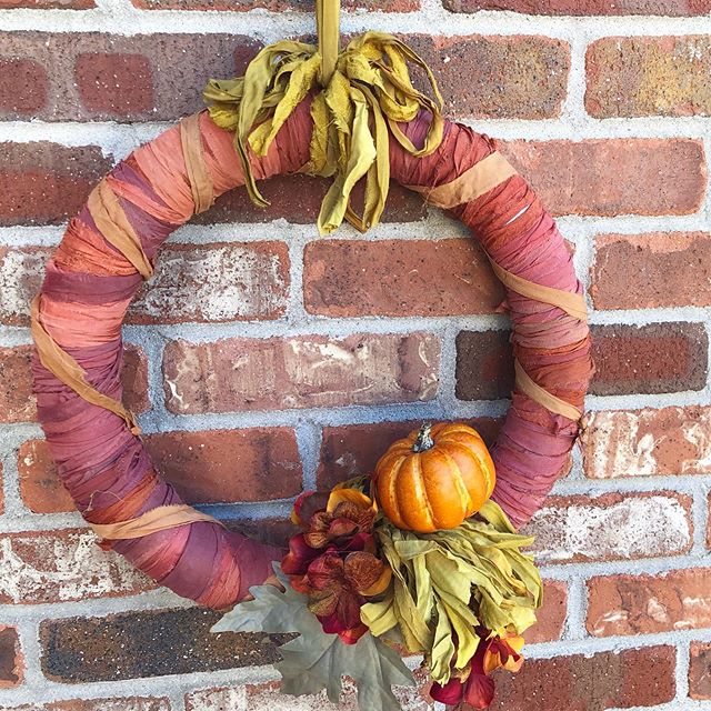 ribbon-wrapped orange fall wreath decorated with fall leaves, pumpkins, and flowers, hung on a brick wall