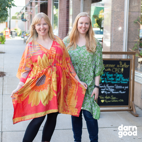 Two women are wearing brightly colored kaftans on the sidewalk of a city.