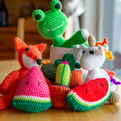A pile of our handmade amigurumi kits are displayed on a wooden table. A frog, unicorn, fox, watermelon, and cactus amigurumis are all completed and posing for the camera!