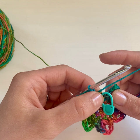 A pair of hands are using a silver crochet hook to crochet the rainbow watercolors lace weight silk yarn, the blue pastel stitch marker attached to their work.