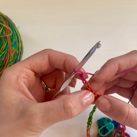 A pair of hands are using a silver crochet hook to crochet the rainbow watercolors lace weight silk yarn into a long chain