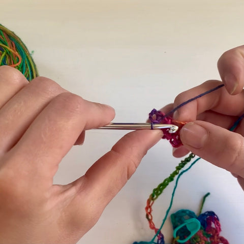 A pair of hands are using a silver crochet hook to crochet the rainbow watercolors lace weight silk yarn into a long chain