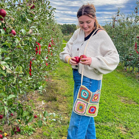 A girl is standing in an apple orchard, putting apples in her granny square crossbody tote bag.