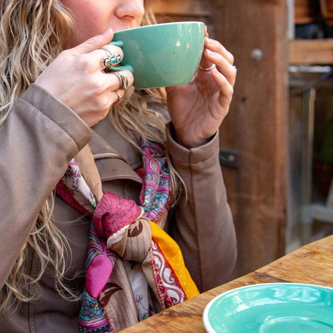 A woman with blonde hair is sitting at a table, wearing a teal cup and a multicolored medley scarf
